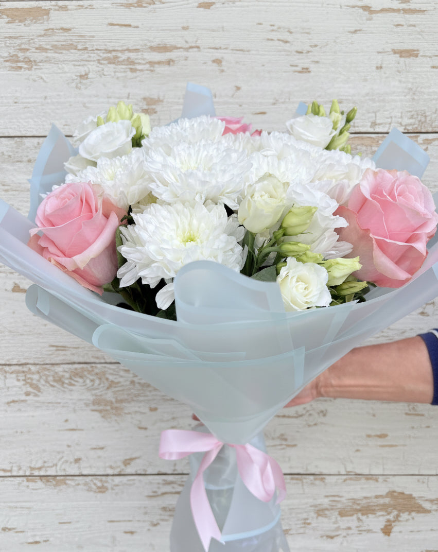 Нежен букет с розови рози, бели хризантеми и еустоми, аранжиран в пастелна опаковка и вързан с розова панделка
