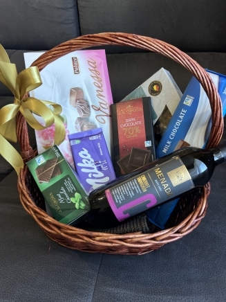 Подаръчна кошница с бутилка вино Menada, шоколад Milka, тъмен шоколад, ментови бонбони и белгийски шоколадови бонбони, украсена със златна панделка.